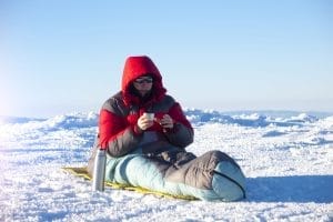 Man som dricker ur termos och sitter i sovsäck på snön.